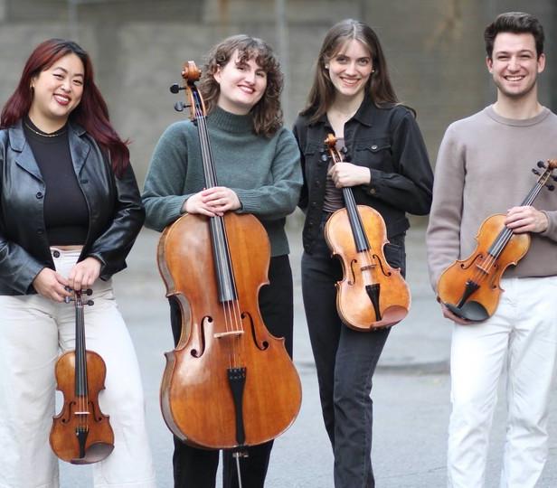 Four musicians stand outdoors holding their string instruments. They are smiling and dressed casually. The group includes two violinists, one violist, and one cellist. They appear relaxed and happy, enjoying each other's company in a casual setting.